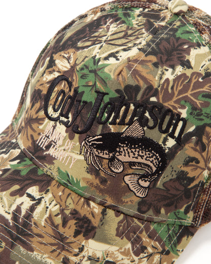 A close-up of a camouflage trucker cap with a leaf and tree bark pattern. The front features a large black embroidered script reading 'Cody Johnson' above a black catfish graphic.