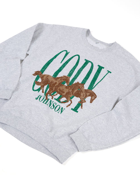 A light heather gray crewneck sweatshirt featuring a graphic print of three brown horses overlaid with green distressed text reading 'CODY' and 'JOHNSON'.