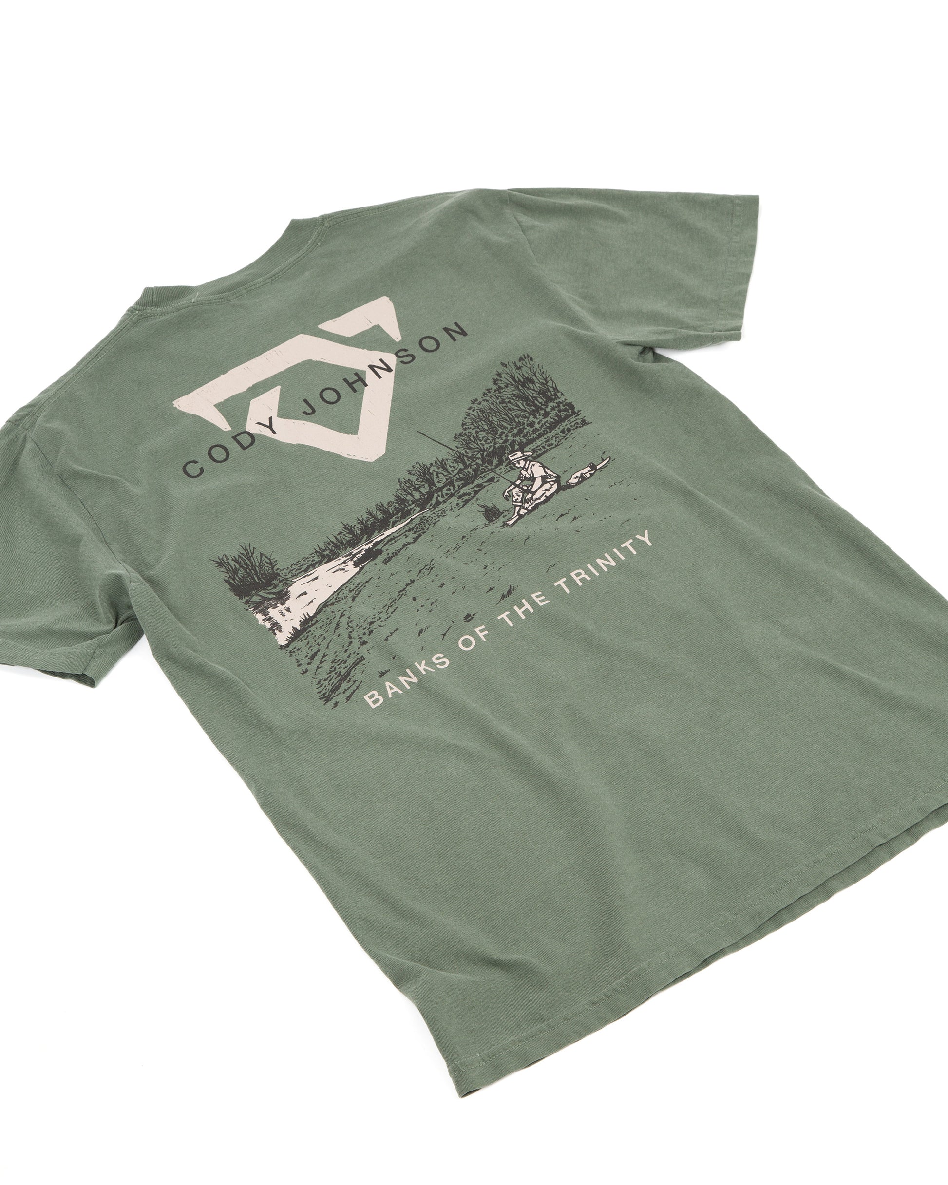 An muted green short-sleeve t-shirt laid, featuring a large graphic print on the back with a COJO bull logo, the text 'CODY JOHNSON', a scenic lake illustration, and 'BANKS OF THE TRINITY' printed below.