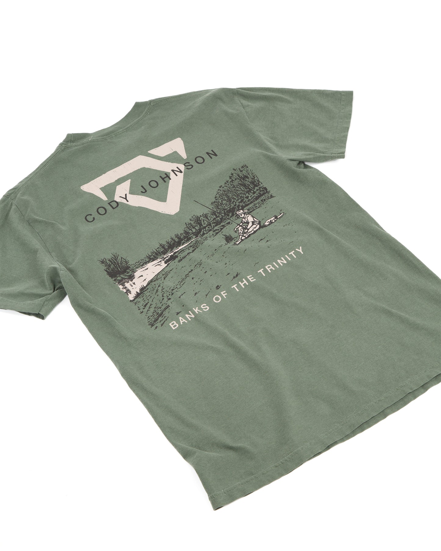 An muted green short-sleeve t-shirt laid, featuring a large graphic print on the back with a COJO bull logo, the text 'CODY JOHNSON', a scenic lake illustration, and 'BANKS OF THE TRINITY' printed below.