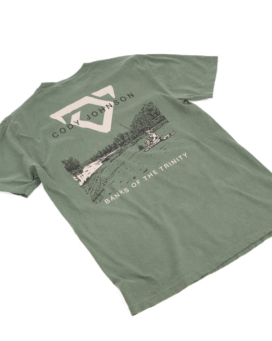 An muted green short-sleeve t-shirt laid, featuring a large graphic print on the back with a COJO bull logo, the text 'CODY JOHNSON', a scenic lake illustration, and 'BANKS OF THE TRINITY' printed below.