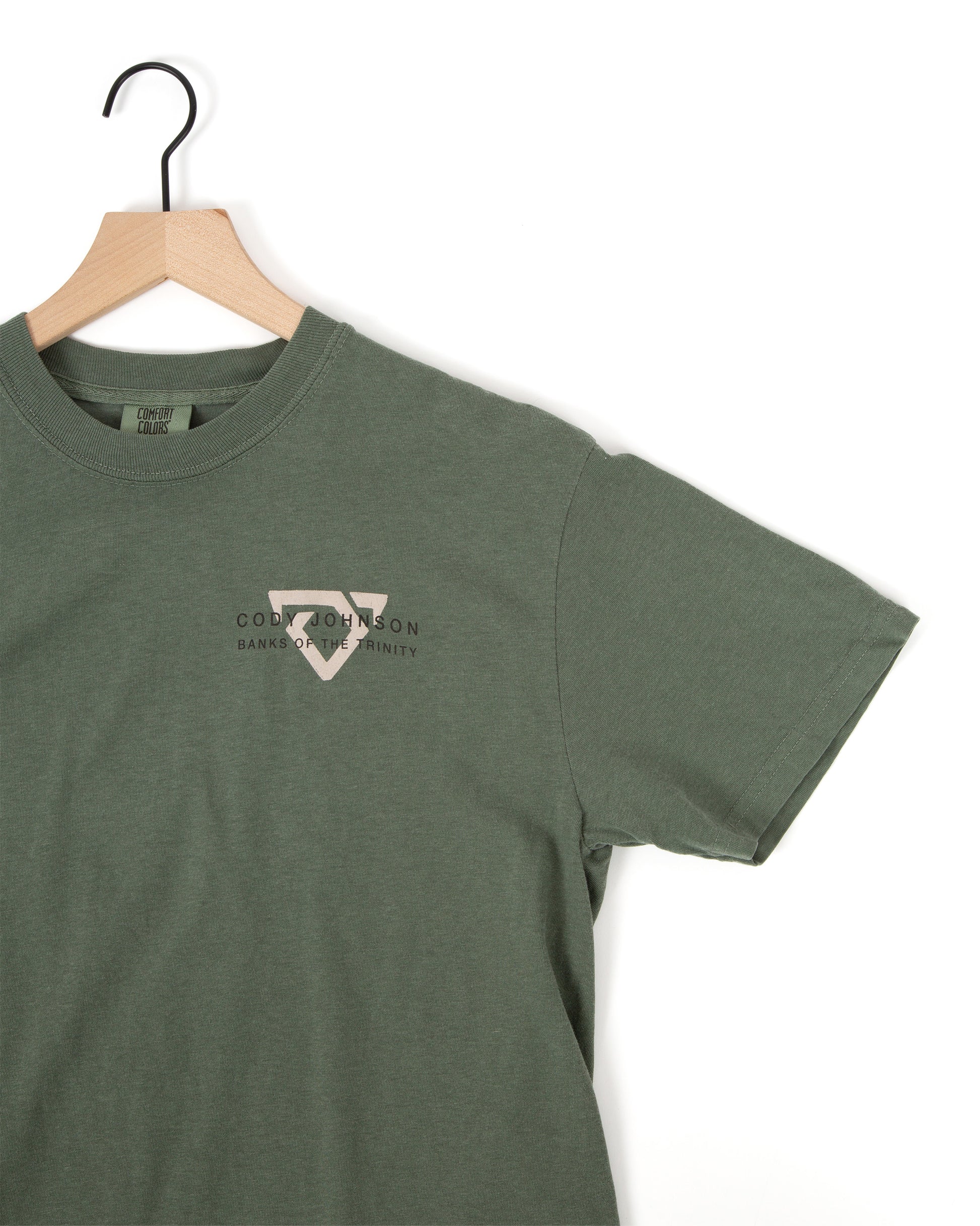 An muted green short-sleeve t-shirt hanging on a wooden hanfer, featuring a small light gray Bull logo on the left chest with the text 'CODY JOHNSON' BANKS OF THE TRINITY