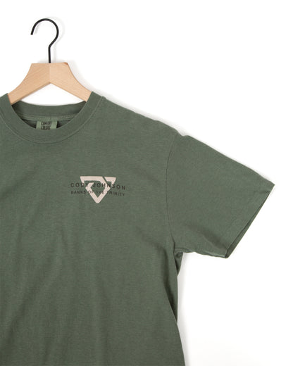 An muted green short-sleeve t-shirt hanging on a wooden hanfer, featuring a small light gray Bull logo on the left chest with the text 'CODY JOHNSON' BANKS OF THE TRINITY