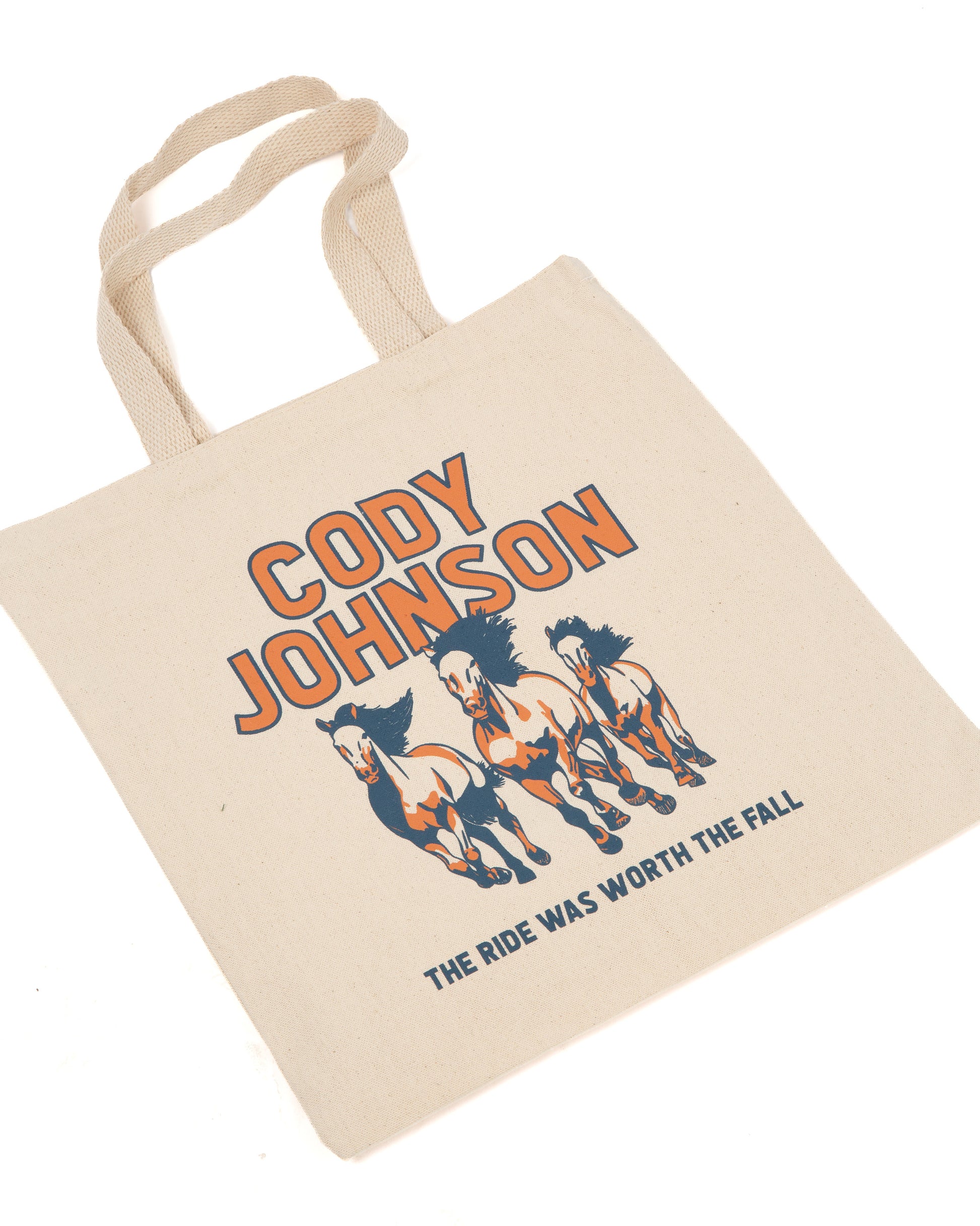 A natural canvas tote bag with printed orange and navy graphics featuring the text 'CODY JOHNSON' and 'THE RIDE WAS WORTH THE FALL' along with three running horses.