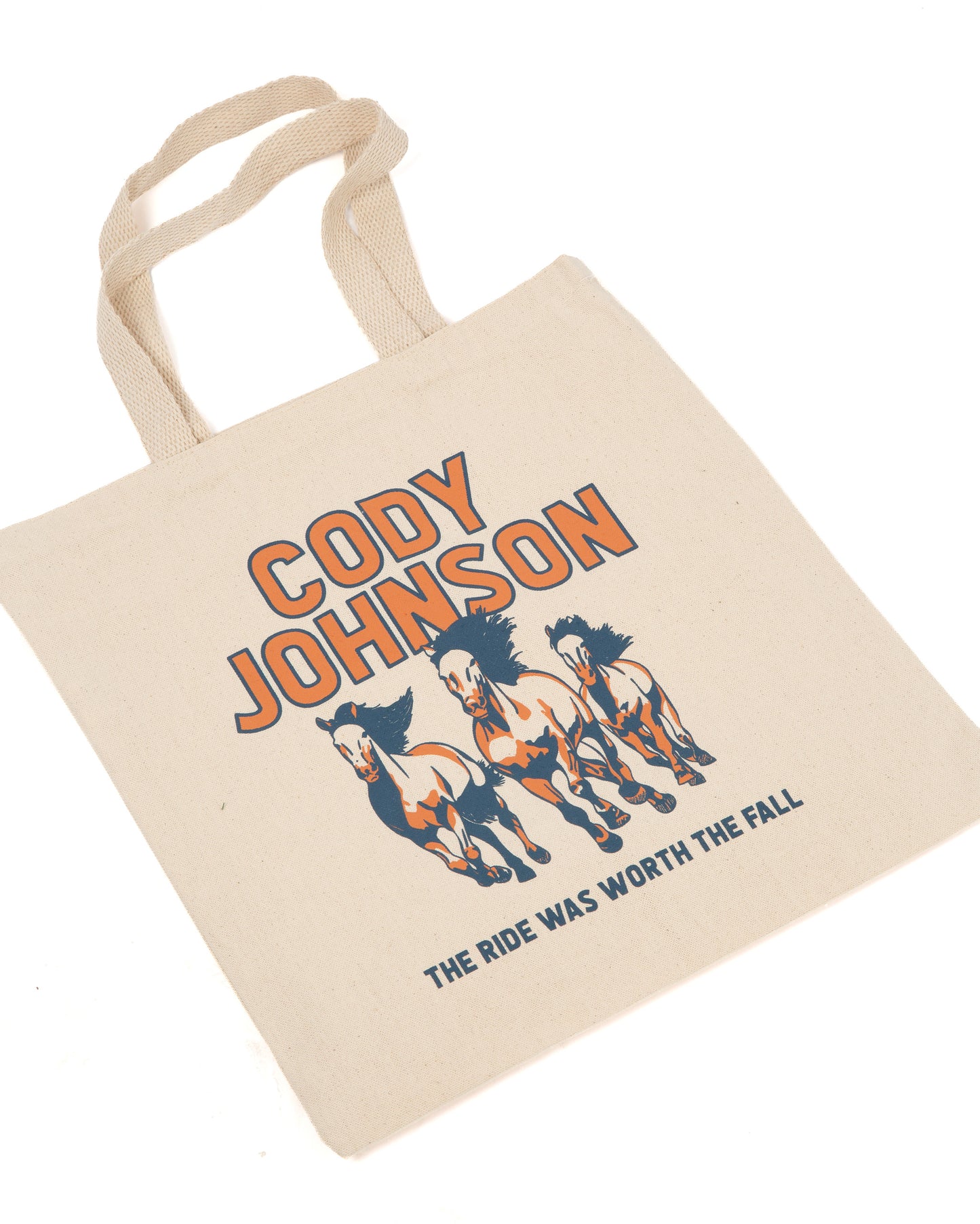 A natural canvas tote bag with printed orange and navy graphics featuring the text 'CODY JOHNSON' and 'THE RIDE WAS WORTH THE FALL' along with three running horses.