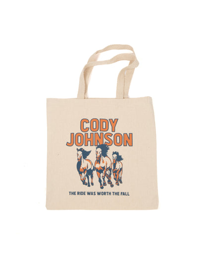 A beige canvas tote bag with a graphic print featuring three horses and the text 'CODY JOHNSON' and 'THE RIDE WAS WORTH THE FALL' in orange and navy blue.