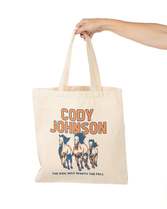 A beige canvas tote bag with a graphic print of four horses and the text 'CODY JOHNSON' and 'THE RIDE WAS WORTH THE FALL', held by a person's arm 