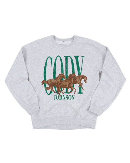 A light heather gray crewneck sweatshirt featuring a graphic print with the word 'CODY' in distressed green letters, three brown horses in the center, and 'JOHNSON' in green text below.